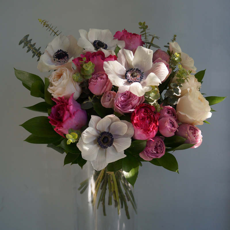 Нежный праздничный букет белых анемонов и розовых роз в прозрачной вазе, украшенный зеленью